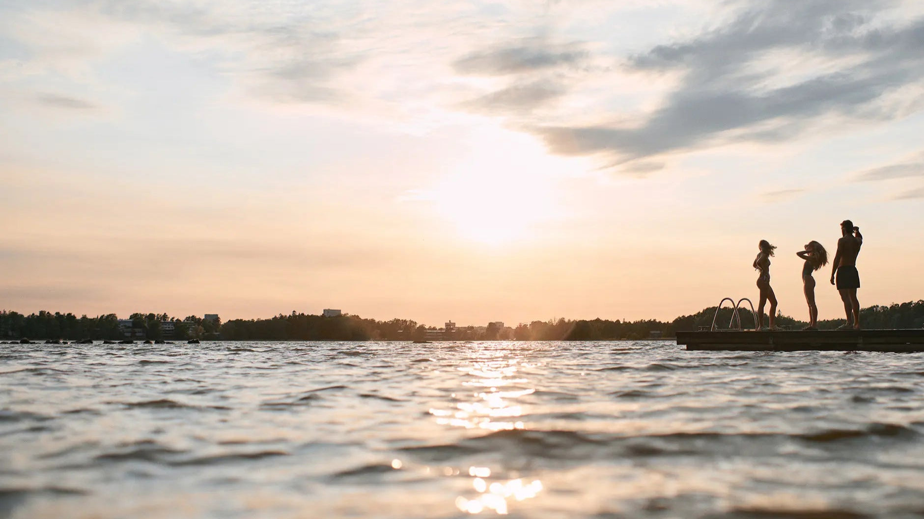 Three people standing on a dock at sunset with water and sky in the foreground wearing ui swim finnish designer swimwear onepieces bikini and swimming shorts.